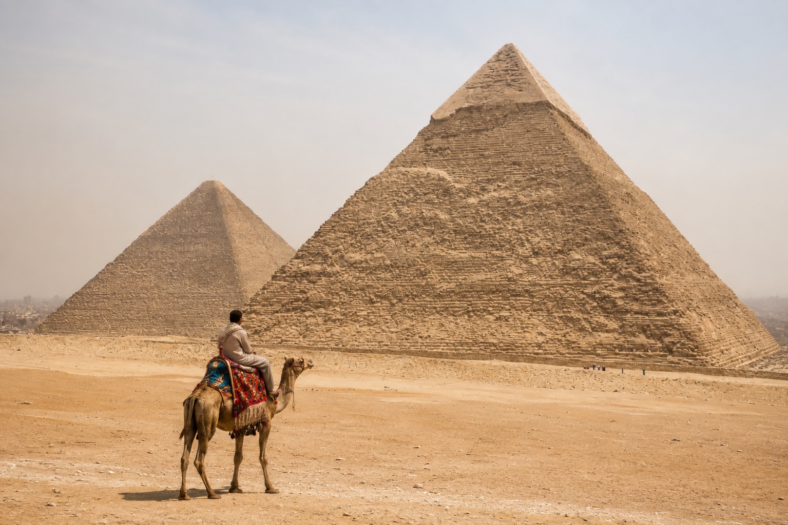Lone camel rider crossing the open sand in daylight before the pyramids of Khafre and Khufu at Giza under a pale hazy sky
