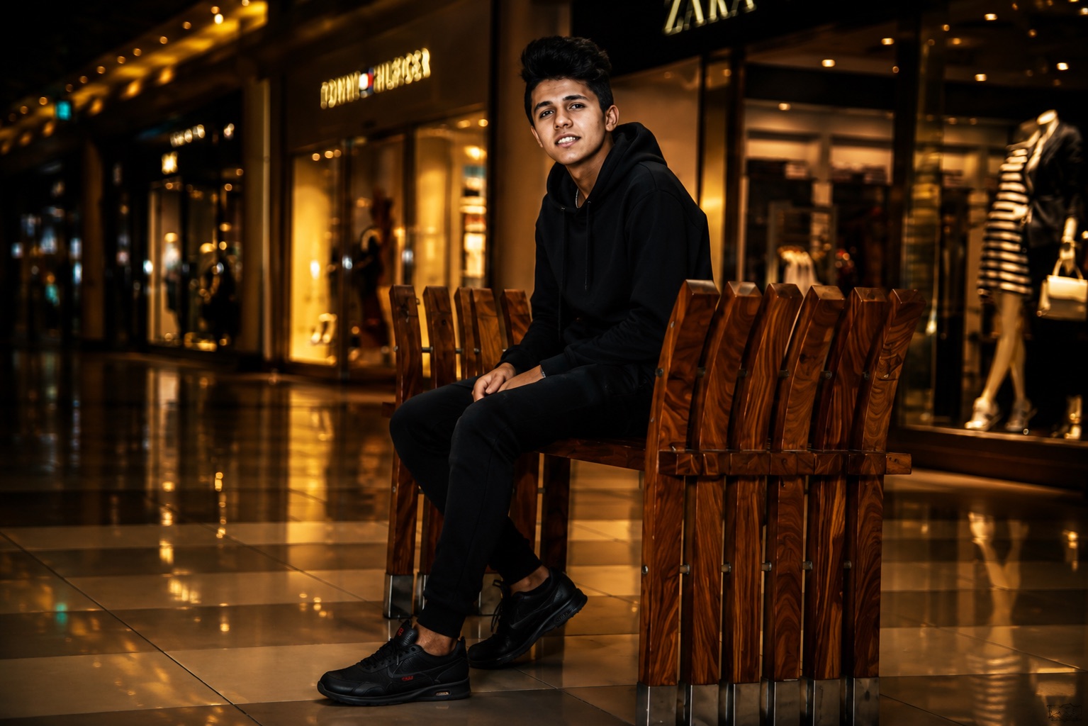 Young man in a black hoodie seated on a bench in a warmly lit shopping mall, shot as an urban lifestyle portrait