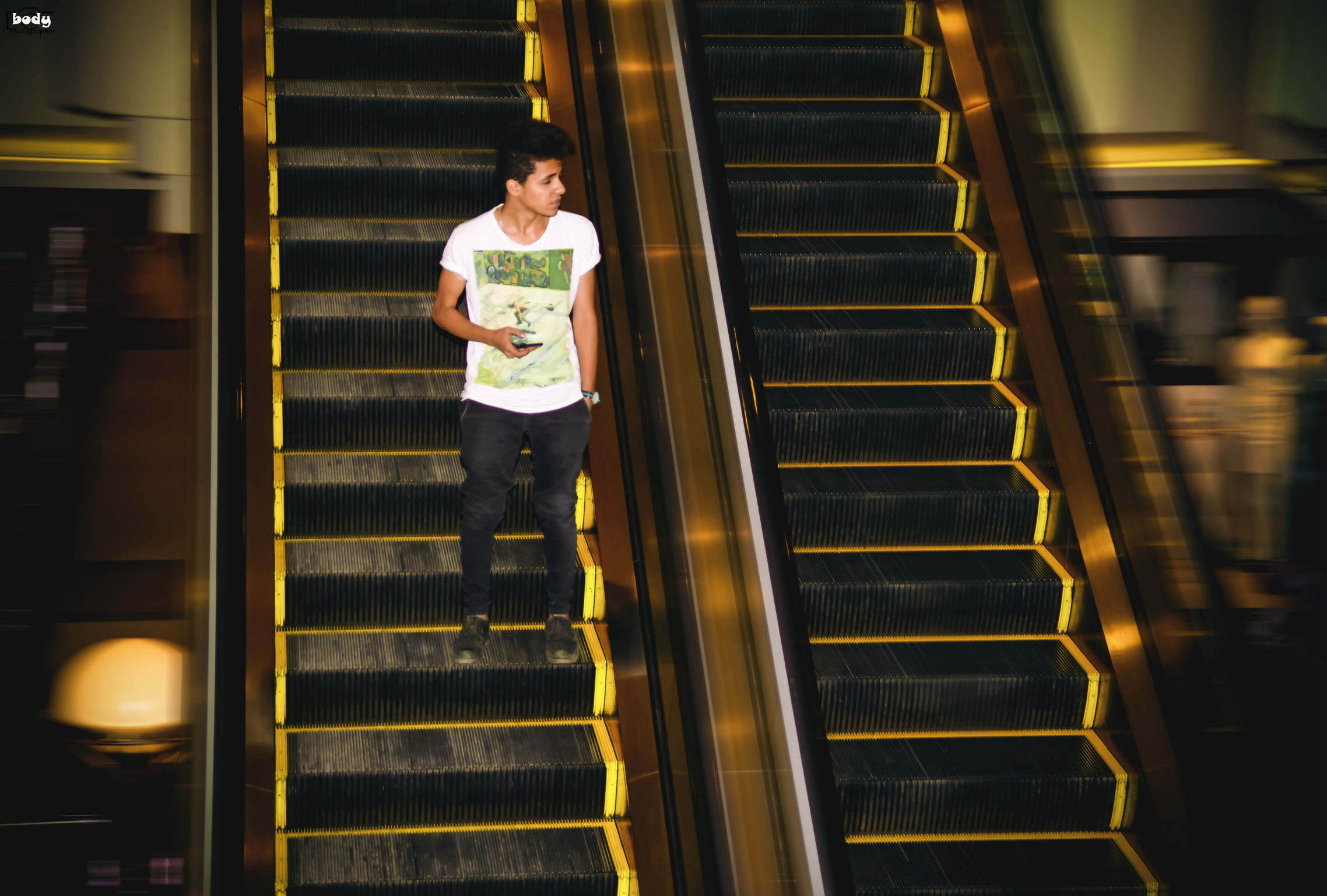 Young man in a white printed t-shirt standing alone on a mall escalator, framed by the yellow-edged steps of two parallel escalators