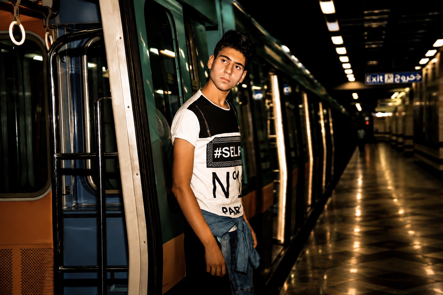 Young man leaning in the open doorway of a metro carriage, the lit platform and exit signs receding down the tunnel behind him
